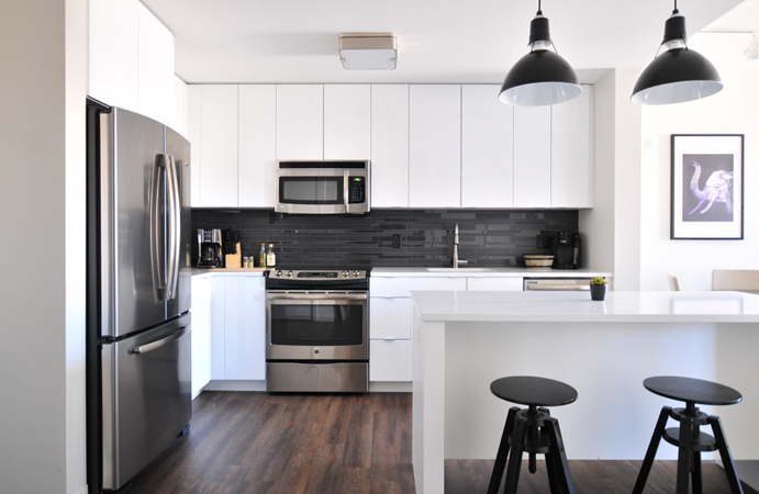 Modern kitchen: white cabinets, dark backsplash, stainless appliances. Black pendant lights over island with stools on wood floor.