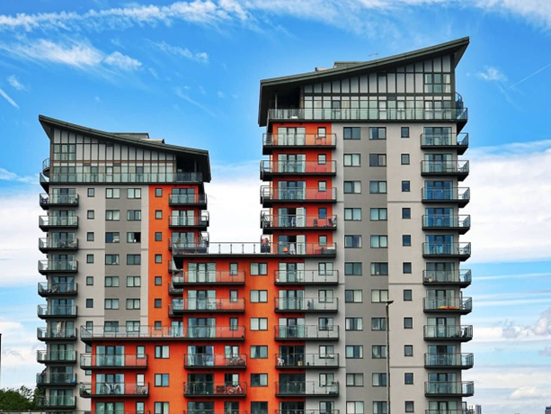 Two modern apartments with angular roofs, red and gray exteriors, lined balconies against a bright blue sky with clouds.