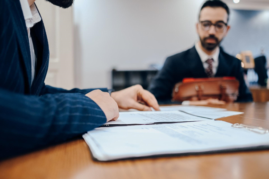 Two men in suits sit at a table with documents; one points to the papers. A leather briefcase hints at a professional setting.