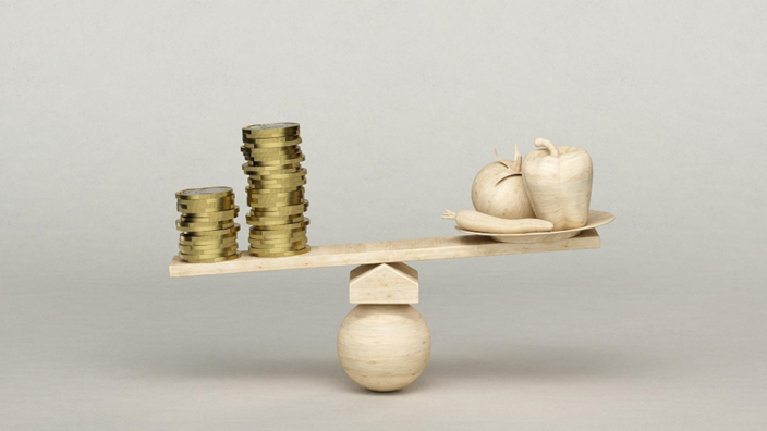 A seesaw on a wooden ball balances gold coins against carved vegetables, illustrating the contrast between money and food.