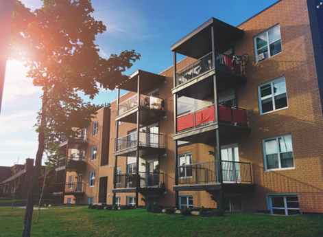 A sunlit four-story brick apartment with balconies, some draped in red fabric. Grass, a tree, and a warm, clear sky are visible.