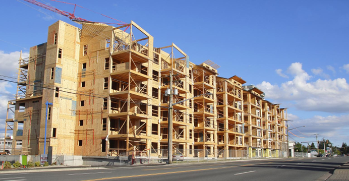 Large wooden building under construction with scaffolding and cranes, set against a blue sky, next to a paved road.