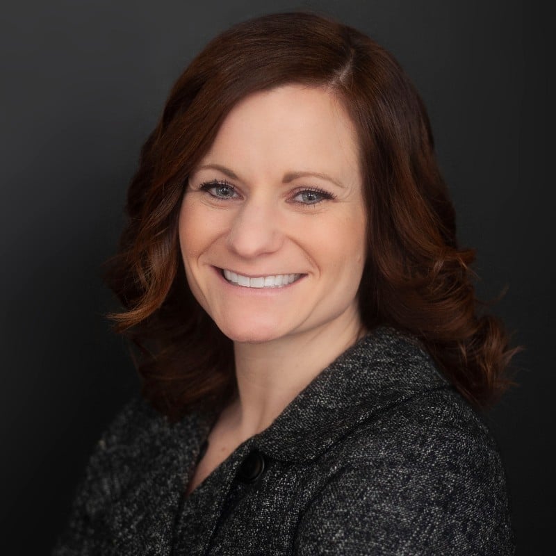 Person with shoulder-length brown hair smiles in a pro headshot, wearing a dark blazer against a dark background.