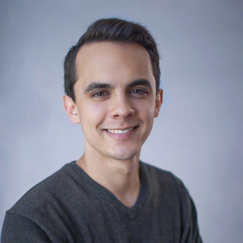 A short-haired person in a dark gray sweater smiles against a light gray background.