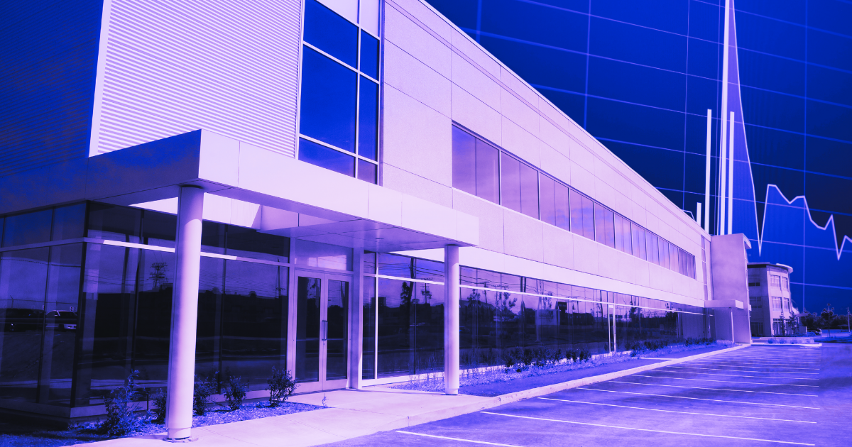 Modern office building with blue-tinted glass facade, sleek lines, columns. Empty parking area in foreground; tall structure behind.