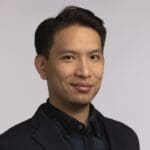 Man with short dark hair in dark blazer poses for headshot on light background, looking composed and slightly smiling.