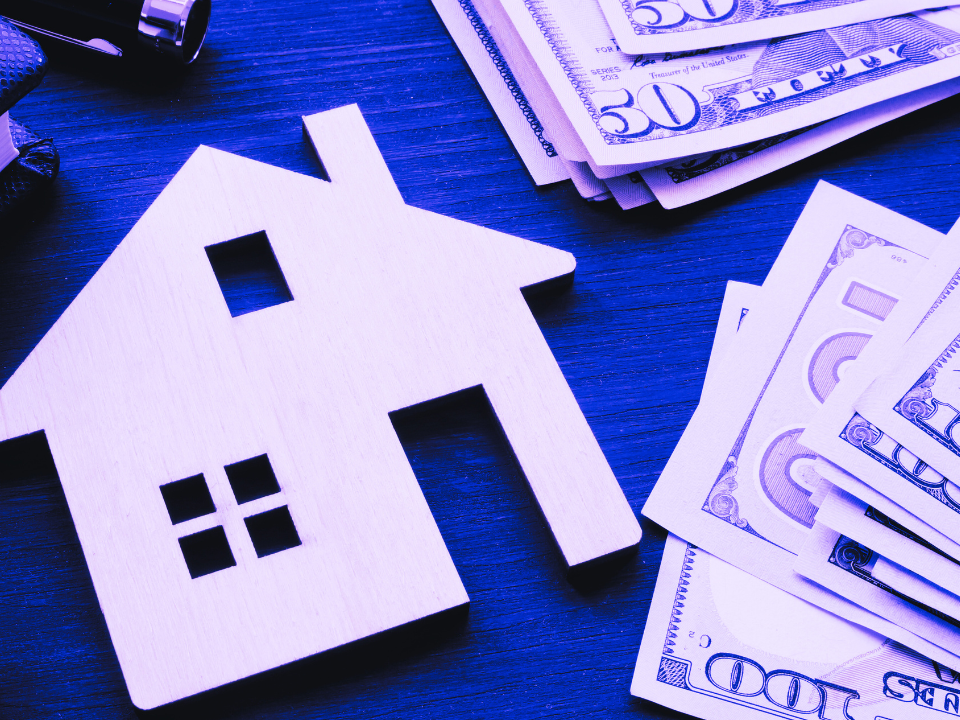 Wooden house cutout and stacked dollar bills on a blue-tinted surface, symbolizing real estate investment or mortgage.