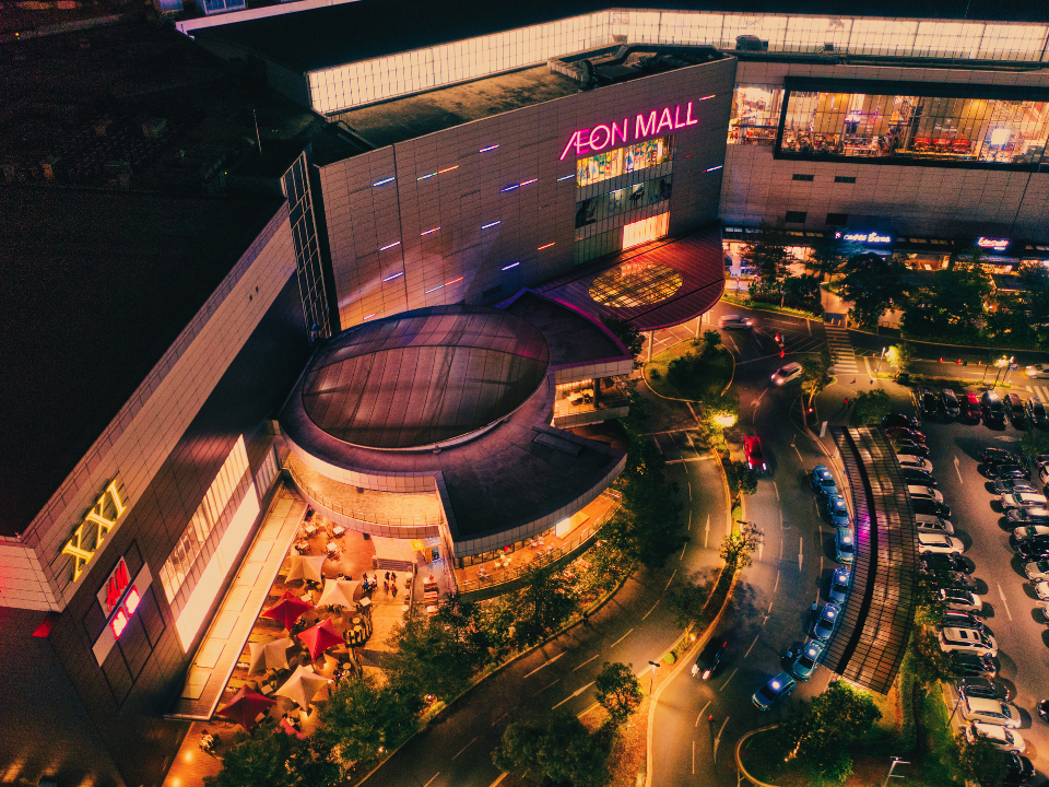 Aerial view of Aeon Mall at night with neon lights, partially filled parking lot, and illuminated trees. Urban nightlife scene.