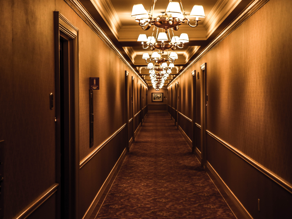 Dim hotel hallway with warm walls and carpet; ornate lights cast a cozy glow. Doors on both sides lead to rooms.