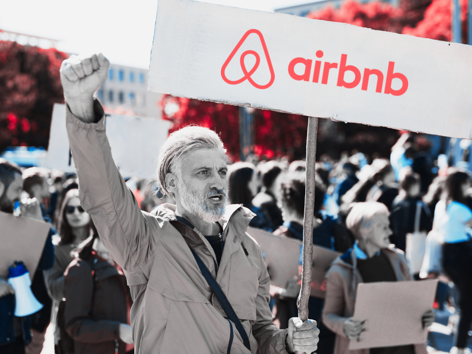 Man holds Airbnb sign during protest, raising fist. Crowd with signs in background. Scene is outdoors for real estate article.