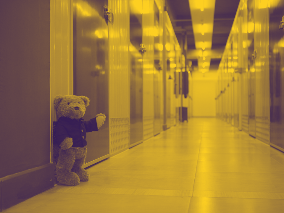 Teddy bear in jacket against a wall in a yellow hallway with closed doors, for real estate article on decreasing self-storage demand.