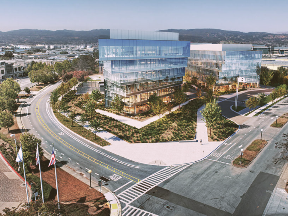 Modern office complex with glass buildings, greenery, crosswalks, flags, and mountains. Featured in an article on life science real estate.