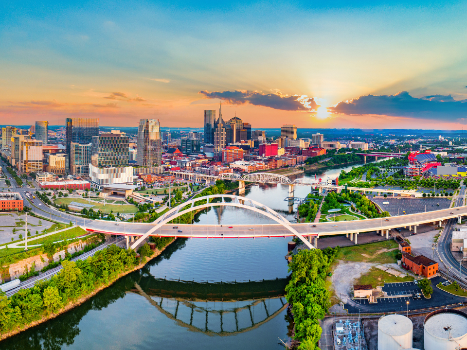 Aerial view of Nashville at sunset, showcasing a white arch bridge over a river, modern skyline, and green riverbanks.