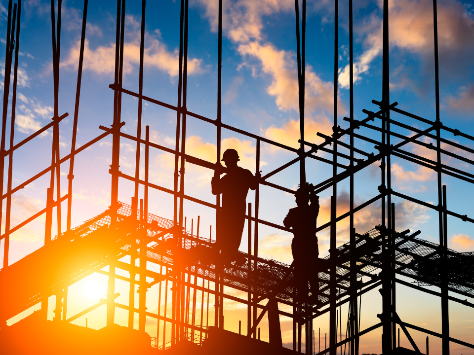 Construction workers silhouetted on scaffolding at sunset, with orange and blue sky. Image for real estate article on builder confidence.