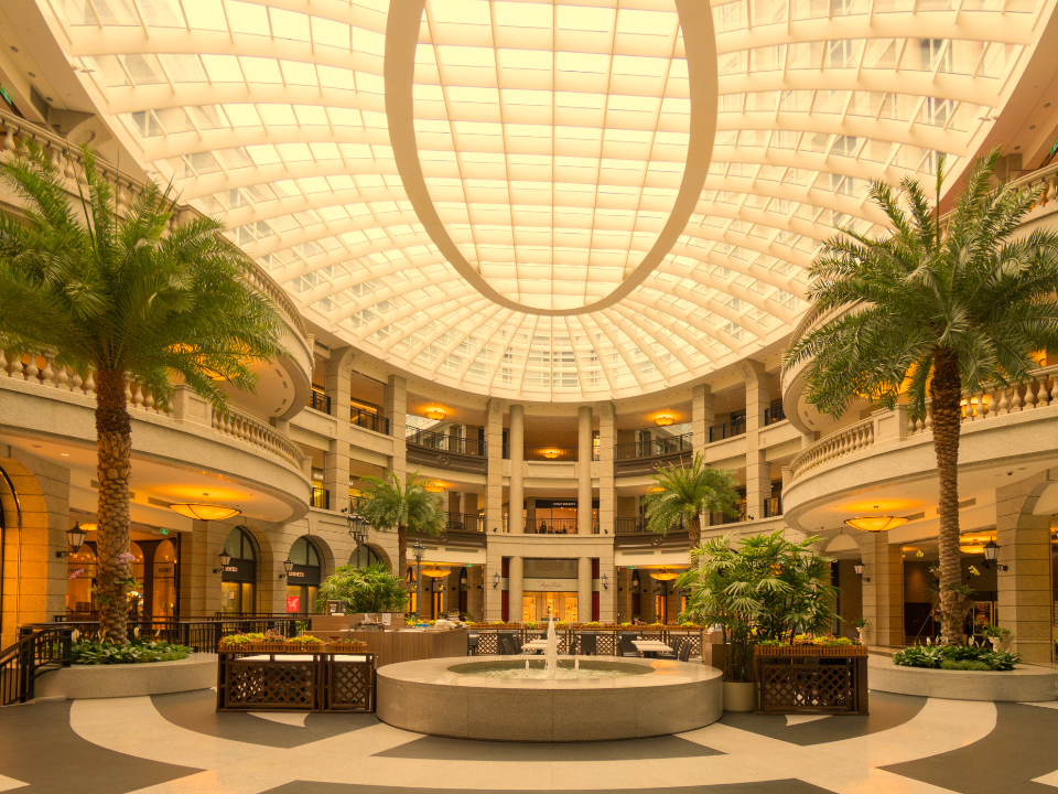 Elegant mall atrium with glass ceiling, palm trees, and central fountain. Multi-level stores under warm lighting.