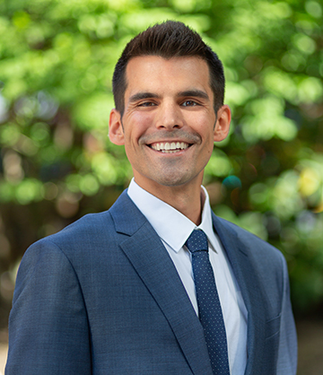 A person in a blue suit and dotted tie smiles warmly at the camera, standing outdoors with green foliage in the background.