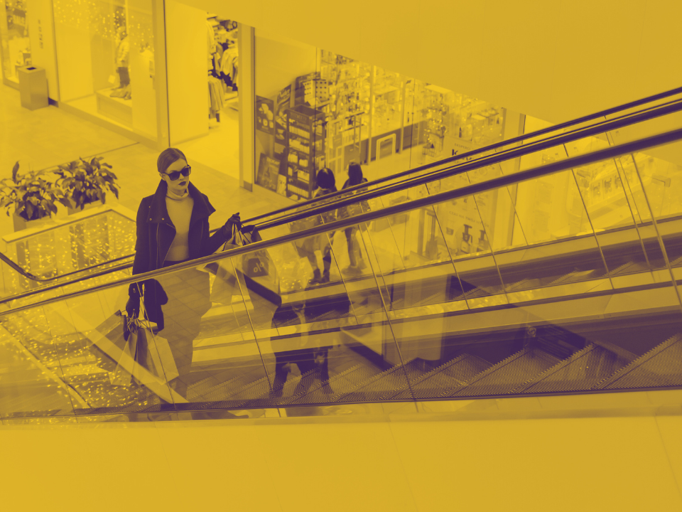 Person in sunglasses with shopping bags rides a mall escalator. Yellow filter; glass panel reflections. For real estate article.