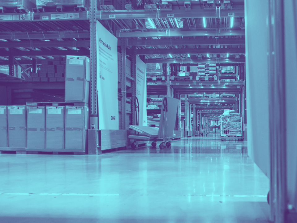 Warehouse interior with tall shelves of boxes, a pallet jack on polished floor reflecting lights; cool, blue tint.