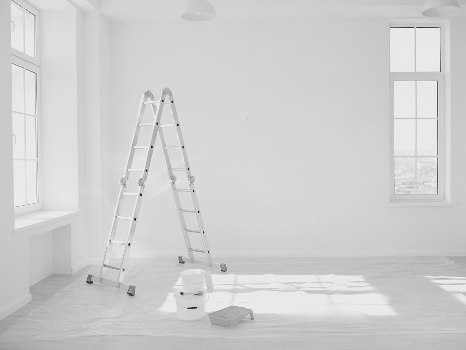 Bright room with high ceiling, ladder on a drop cloth, paint supplies nearby. Large windows illuminate clean white walls.