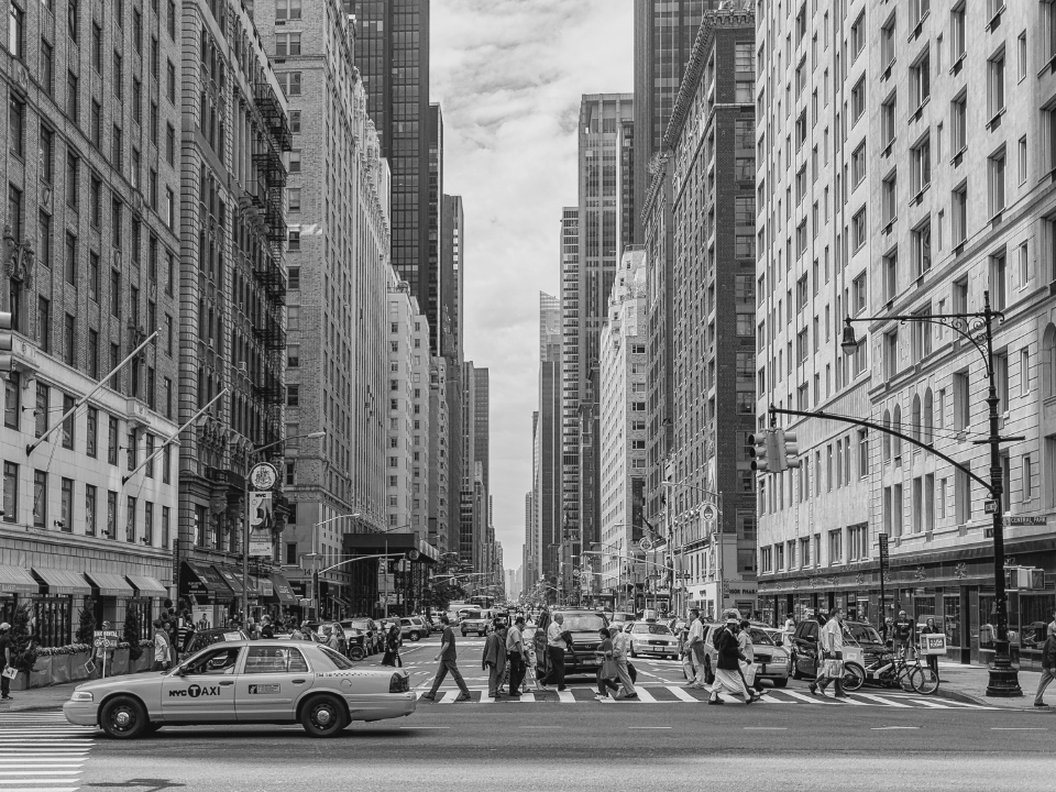 Black and white city street with people, cars, and buildings. Urban life scene for article: "NYC Apartment Construction Slows.