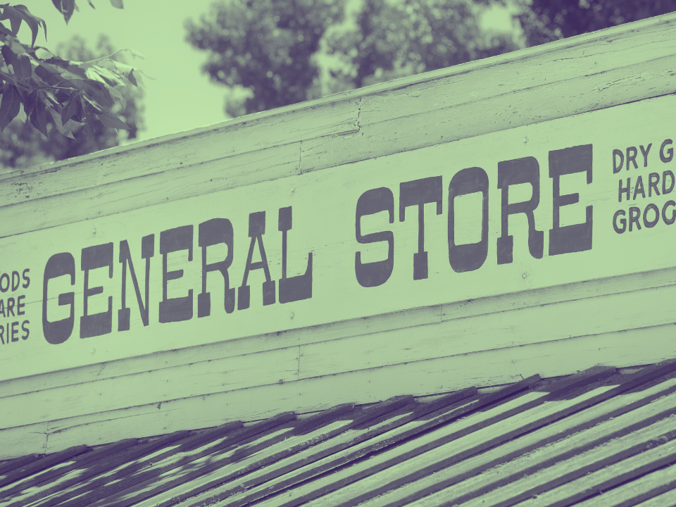 General store sign with vintage lettering: Dry Goods, Hardware, Groceries. Trees and bright sky in the background.