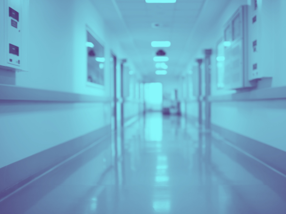 Empty hospital corridor with bright lights, polished floors, white walls, notice boards, and equipment. Featured in HUD article.