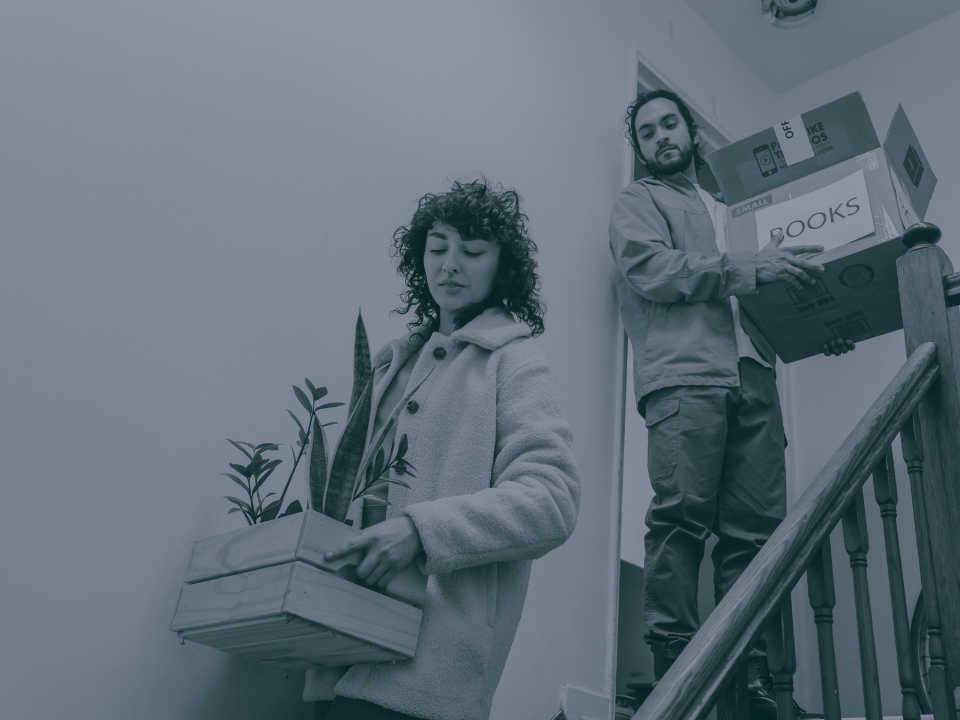 Person in coat carrying plant box down stairs, followed by someone with a box labeled BOOKS; indoor scene with a blue tint.