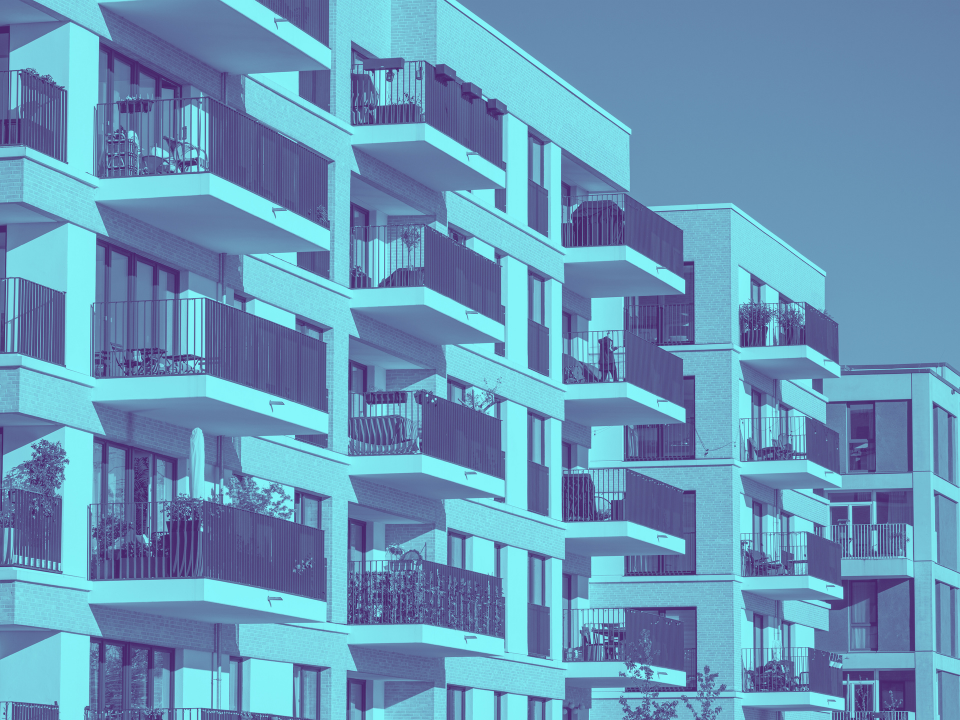 Modern apartment building with balconies, blue tint. Outdoor furniture and plants visible. Clear sky adds a serene atmosphere.