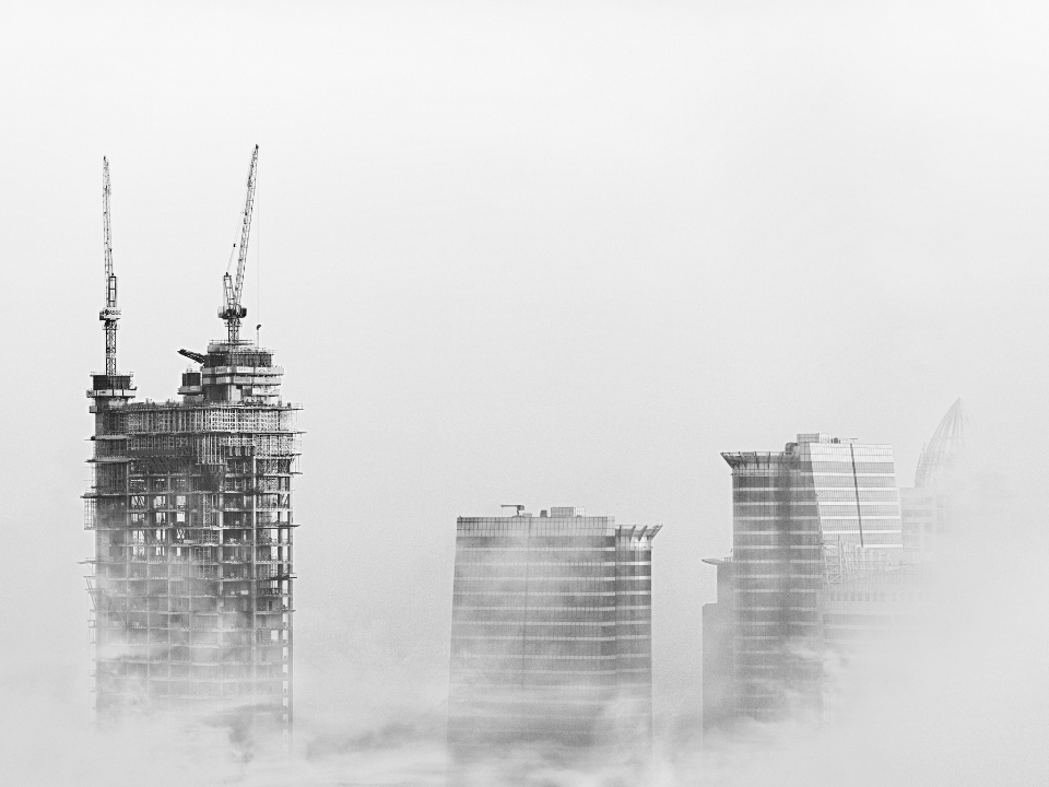 Foggy black-and-white photo of skyscrapers and cranes atop a building under construction, showcasing a modern urban cityscape.