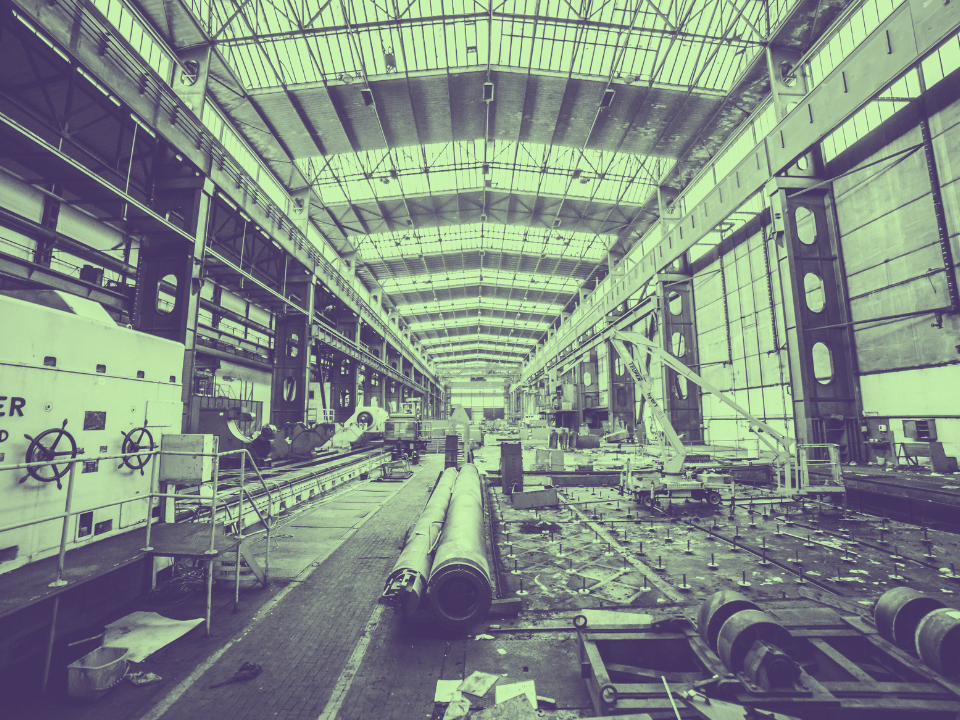 Industrial warehouse with skylights, machinery, and metal beams; view down aisle with open entrance. Real estate article image.