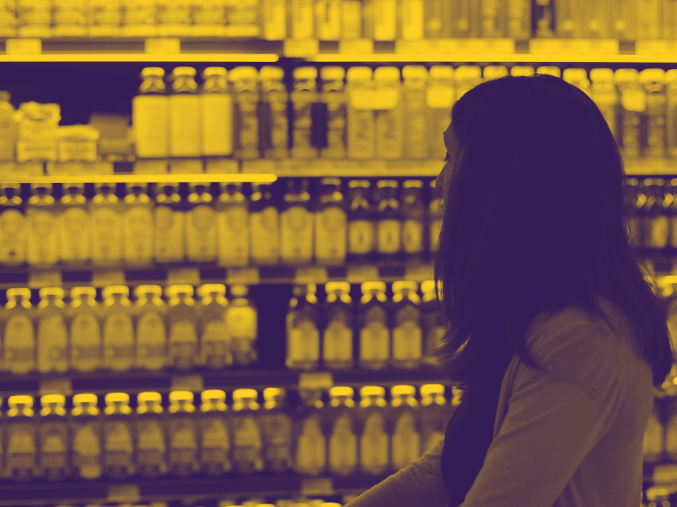 Woman with long hair in a grocery aisle, examining products on a yellow-hued shelf, for an article titled "Kroger and Albertsons.