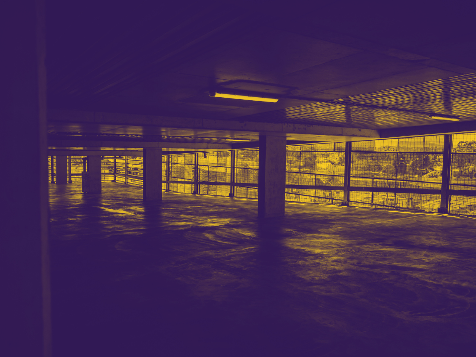 Dim, yellow-tinted view of an empty parking garage. Urban buildings visible through railings. For a real estate website article.